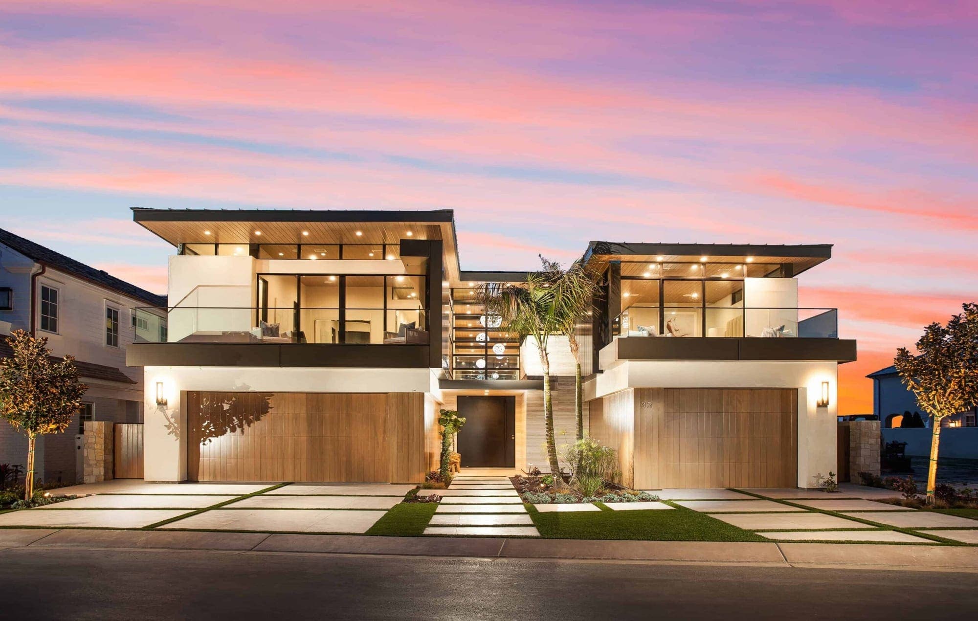 Beach View II, a finished Genova home in Newport Beach under a pink sunset sky, warm wood and lit glass across both upper floors.