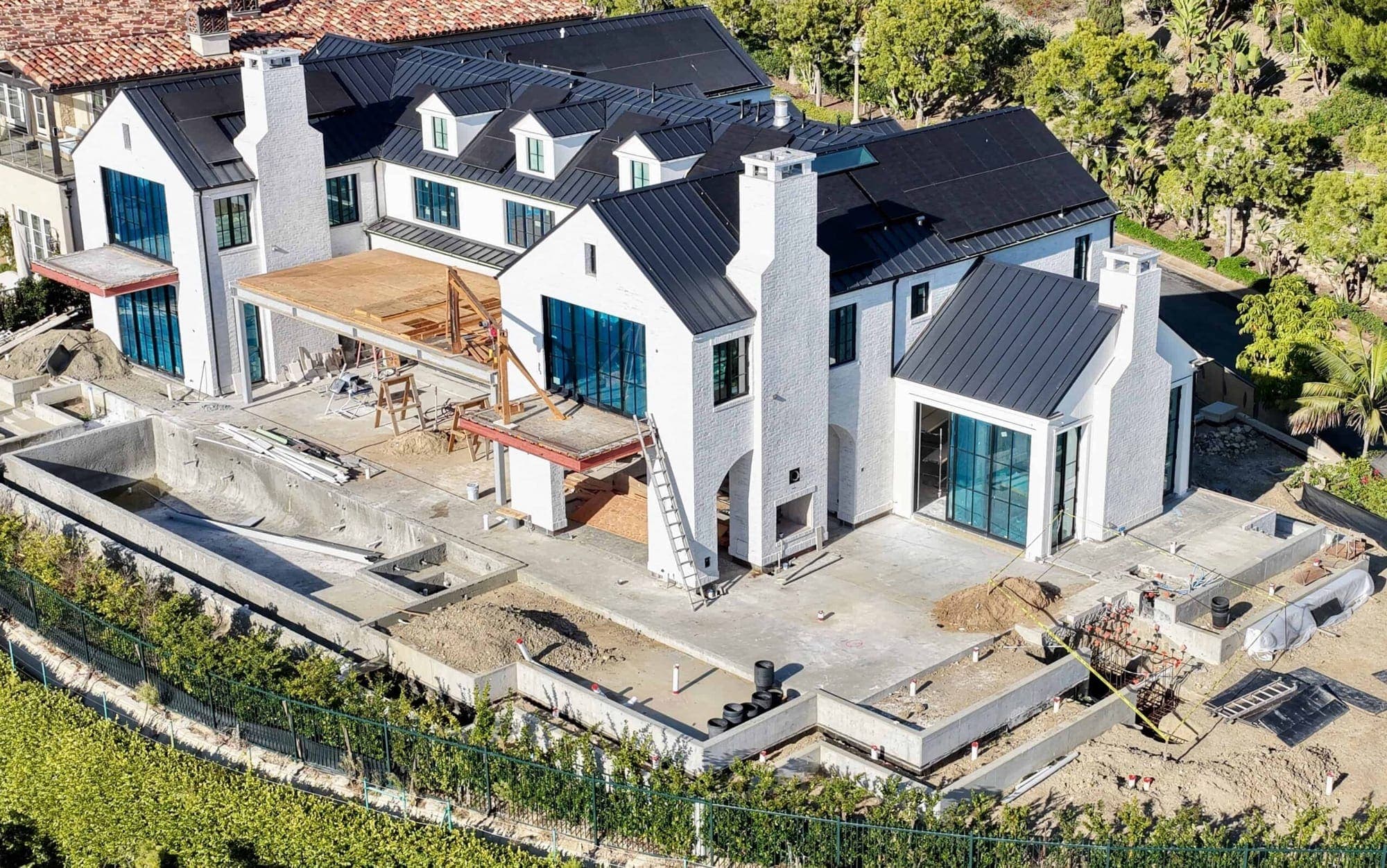 An aerial view of a white brick Dana Point home under construction, black metal roof on, terraces poured, and the pool still an open shell.