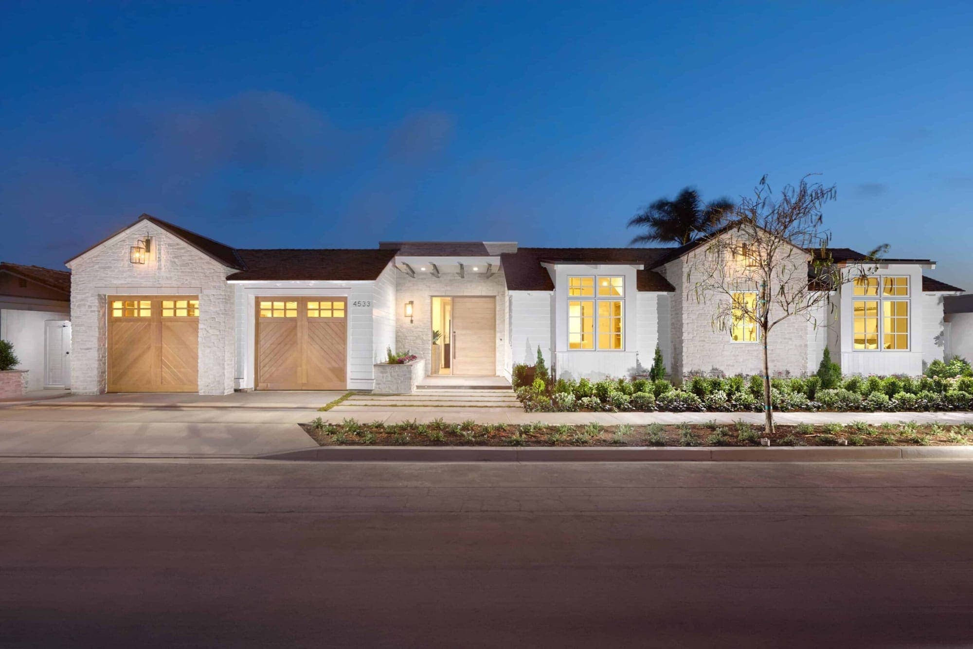 A single-story white Corona del Mar home at dusk, its oak garage doors and windows lit from within.