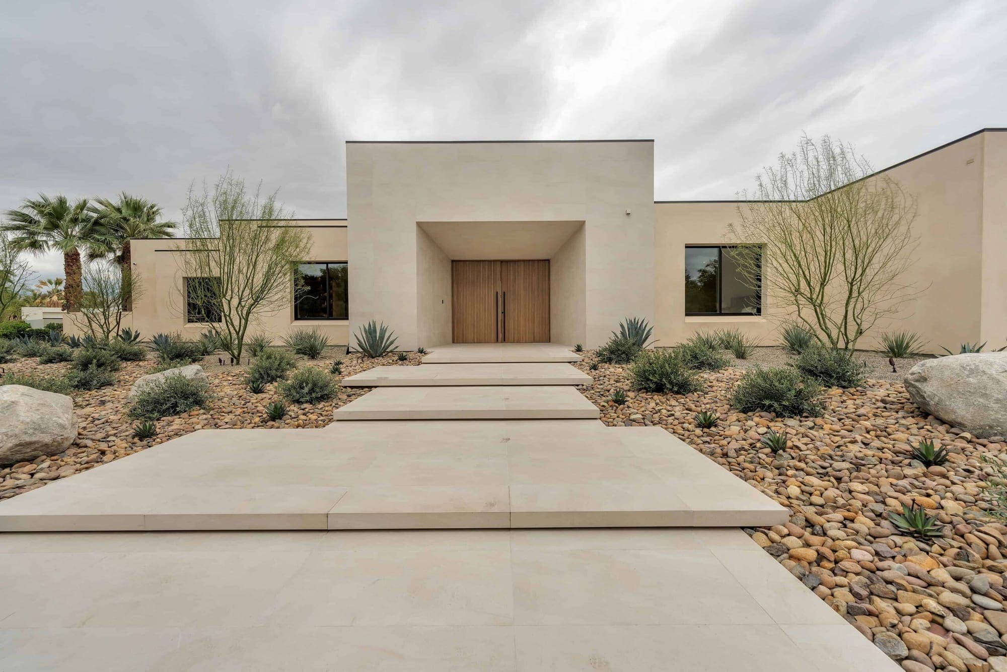 A single-story desert-modern home with a tall stucco entry, rift-oak doors, and rock and agave landscaping.
