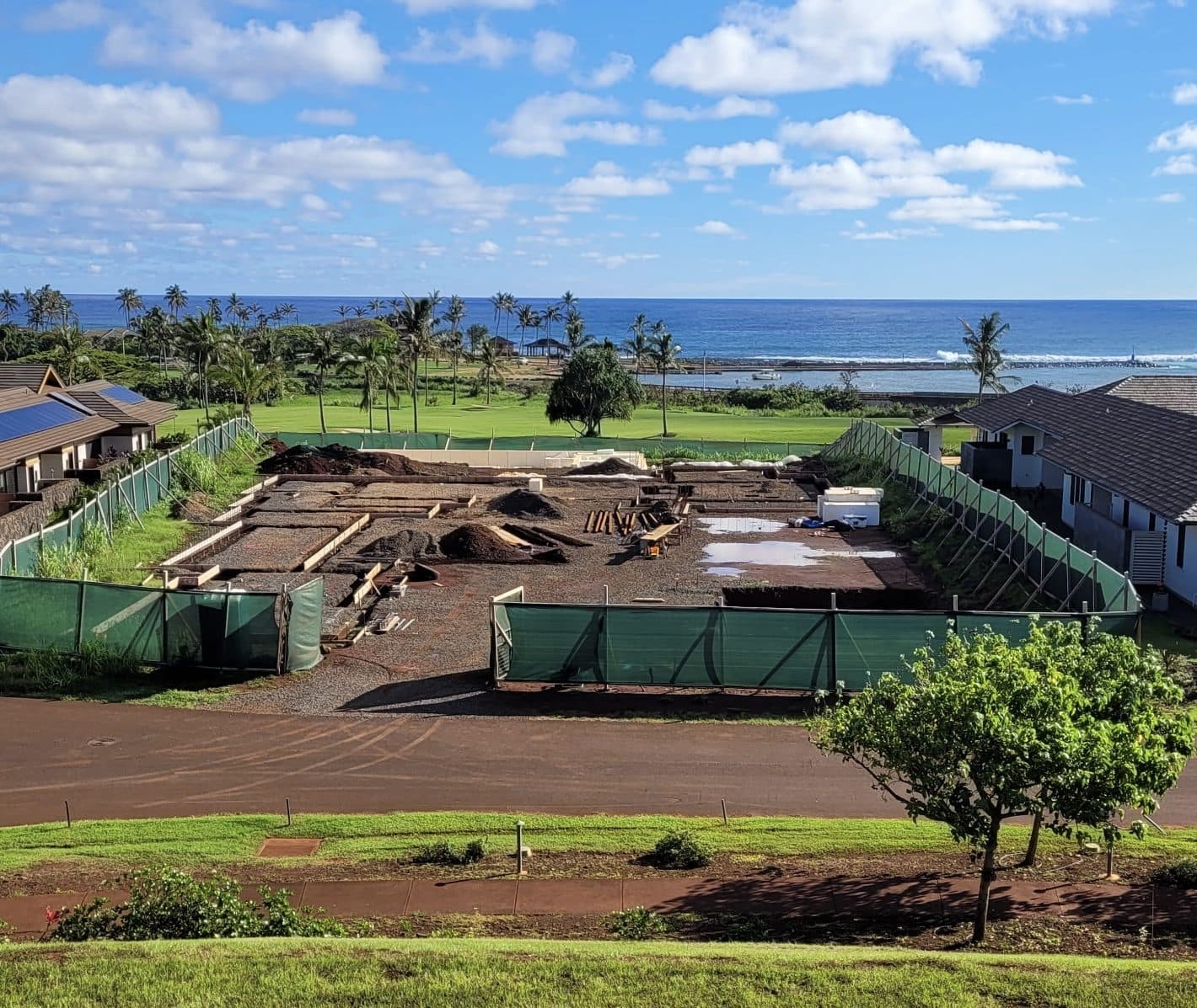 A Genova home site under construction in Kukui’ula on Kauai, the foundation formed and fenced, with palms, a green fairway and the Pacific beyond.