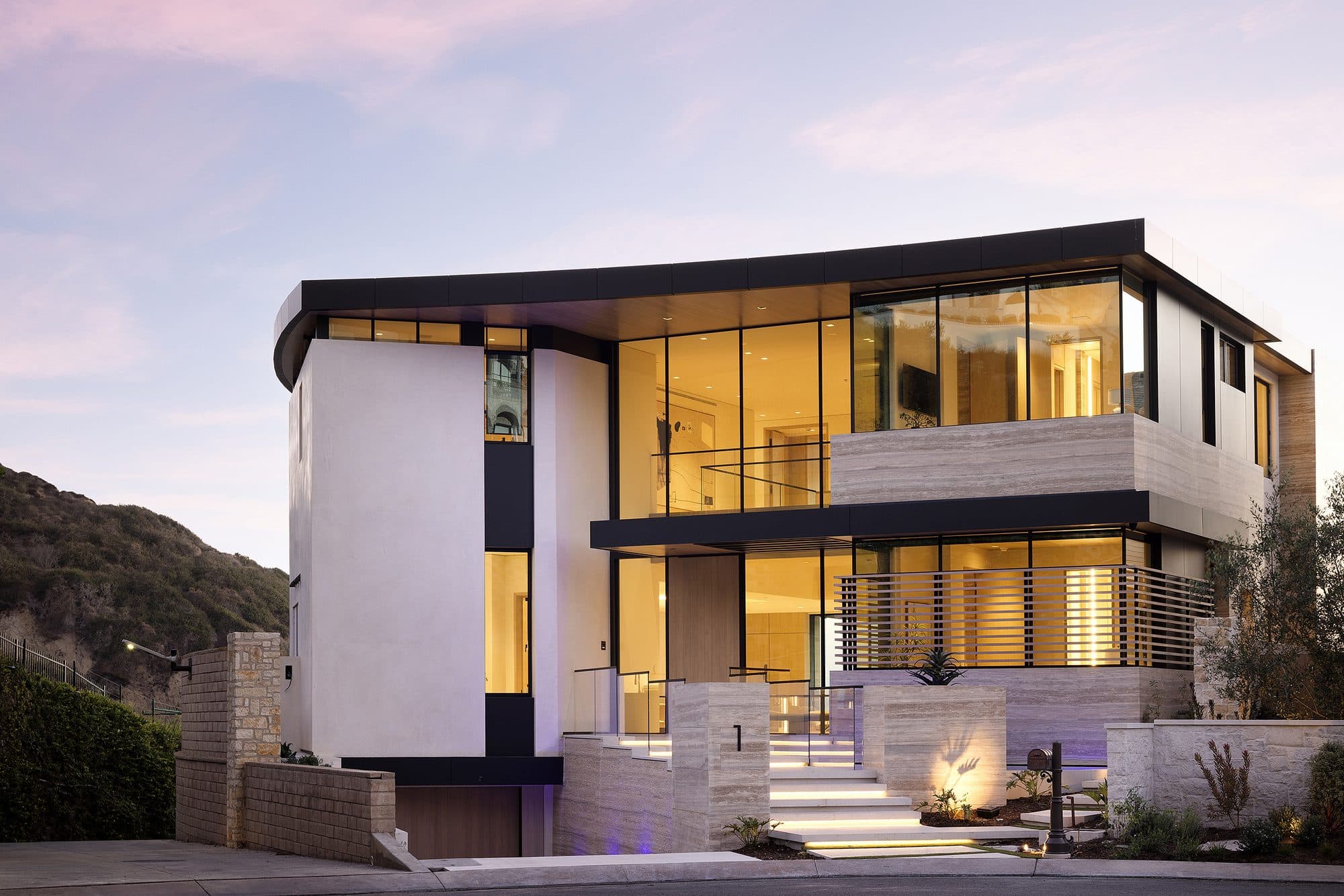 A contemporary white and glass home at The Strand in Dana Point at dusk, its upper floors cantilevered over a lit entry with the headland behind.