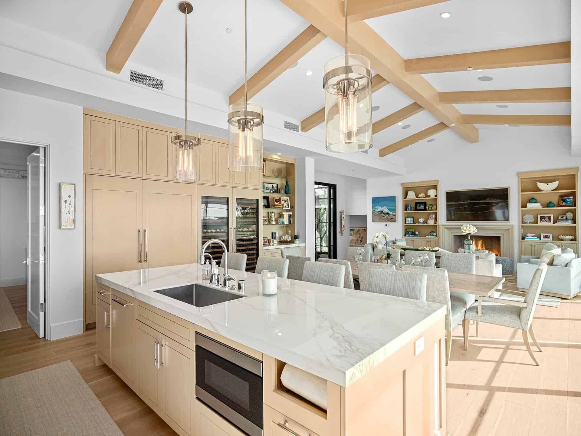 A bright, open coastal kitchen with a marble island, wood beams and a wall of glass to the outdoors.